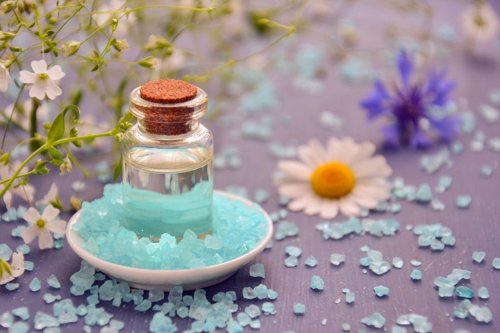 Petit flacon d’huile essentielle posé sur des sels de bain bleus avec des fleurs autour.